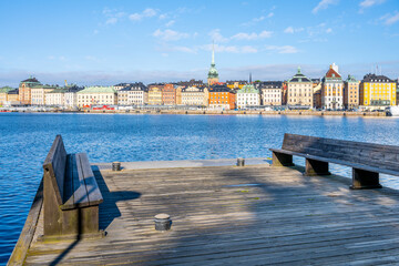 A panoramic view from a wooden pier showcases the vibrant facades of Stockholm Old Town, reflecting in the tranquil waters under a clear blue sky. Sweden