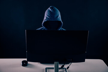 Anonymous and faceless hacker under hoodie using computer isolated over dark background - illegal...