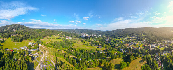 An aerial view reveals the sprawling landscape of Tanvald, stitching together dense green forests, a network of roads, and clusters of buildings under a clear sky. Jizera Mountains, Czech Republic