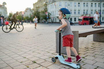 Adorable toddler boy riding his scooter in a city on sunny summer evening. Young child riding a roller.