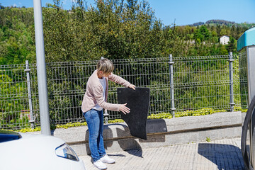 A woman vigorously shakes a car mat, cleaning it at a service station.