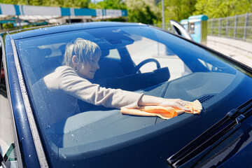 Cheerful woman wipes her car's windshield with a cloth under the sun.