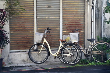 This photo depicts a daytime scene in an alley with two parked bicycles. The house behind them is...
