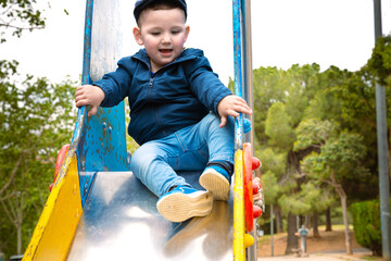  Sweet little Caucasian boy in cap having fun smiling widely sliding down the slide. - obrazy, fototapety, plakaty