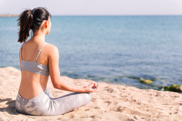 Fototapeta na wymiar unrecognizable young woman in sportswear doing meditation at beach sitting with legs crossed, concept of mental relaxation and healthy lifestyle