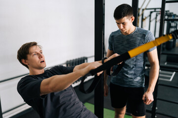 Male using looped ropes doing all-body resistance training. Beginner sportsman doing fitness trx exercises in gym with personal fitness trainer. Concept of healthy lifestyle, physical sport activity.