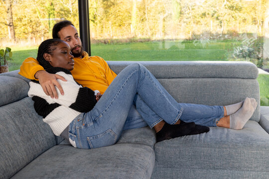 A multiracial couple enjoys a quiet, intimate moment together, reclining comfortably on a gray sofa. They are in a peaceful embrace, dressed casually in denim and soft sweaters, with a bright, natural