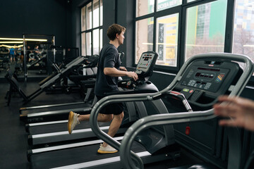Fototapeta na wymiar Rear remote view of athletic man jogging on gym machine, having cardio session in well-equipped fitness club. Side view of beginner sportsman having intensive cardio workout to burn calories in gym.