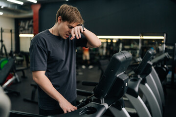 Portrait of tired beginner runner sportsman resting after running on treadmill during cardio session in gym. Satisfied fit male standing on treadmill in fitness gym, feeling exhausted after jogging.