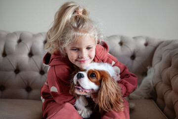 Beautiful blond girl child sits at home on a sofa, playing with and hugging dog, creating heartwarming moments of joy and companionship with pet - obrazy, fototapety, plakaty