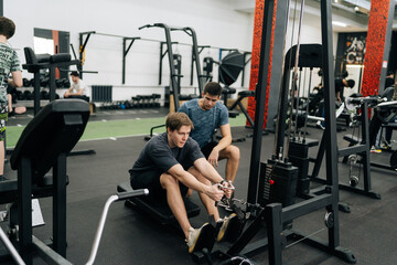 Remote wide shot of beginner sportsman with personal instructor working out in gym by using rowing machine. Sweating man doing exercises for back muscles under supervision of trainer measuring time