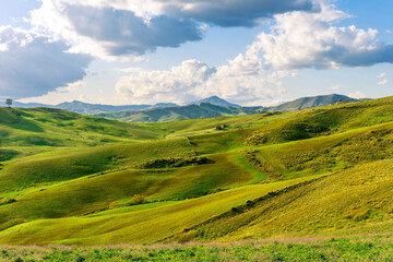 beautiful rural view at summer or spring season fields and hills with rustic grassland and nice mountains with blue cloudy sky on background of countyside landscape