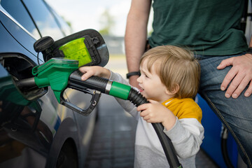 Cute little blond boy holding pump nozzle. Small funny kid helping father to fuel the car at a gas station. - 794970866