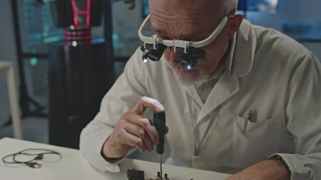 Waist up of mature Caucasian male engineer in magnifying glasses with light using screwdriver while repairing motherboard at work desk in dark engineering workshop