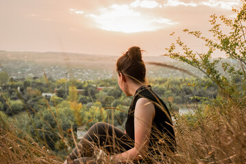 Woman sitting on hillside, enjoying a tranquil sunset over a countryside landscape, reflecting on...