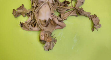 Young sexy woman with blond hair artfully covered with dry, withered decorative banana tree lies on the green studio floor