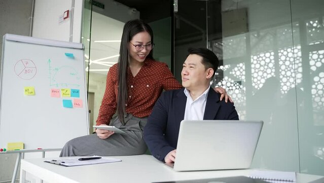 Young asian female employee sexually harasses male co-worker at workplace in business office. Attractive beautiful woman sits down at desk and puts her hand on shoulder of shocked displeased colleague