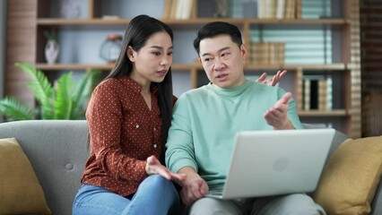 Asian family couple arguing while shopping online using laptop while sitting on sofa in living room at home. They are looking for a product in online store. A husband is unhappy with his wife's choice