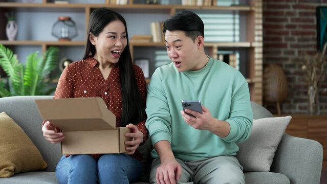 Joyful asian family couple customer opens package at home. Happy wife and husband buyer unpacking purchase sitting on sofa. Satisfied client of e-commerce. Good feedback. Express delivery services 
