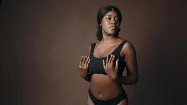 Portrait of young beautiful Black woman in underwear with navel piercing touching her skin gently and posing for camera in studio