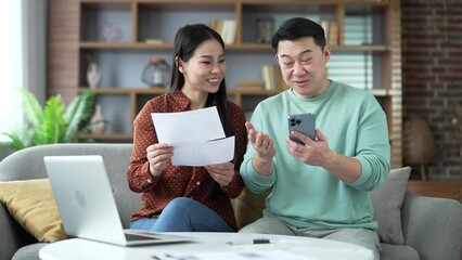 Happy asian married couple dealing with family finances using website or phone app while sitting at home. Smiling wife is holding a utility bill in her hands, and her husband is making payments online