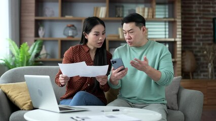 Frustrated shocked asian married couple looking at large utility bills using phone sitting in home. Upset woman holding a paycheck. Confused husband and wife having difficulty doing household finances - Powered by Adobe