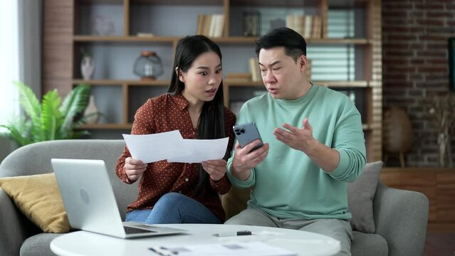 Frustrated shocked asian married couple looking at large utility bills using phone sitting in home. Upset woman holding a paycheck. Confused husband and wife having difficulty doing household finances