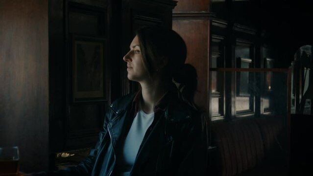 Young woman sitting in a pub, with a drink. Looking out the window, cinematic.