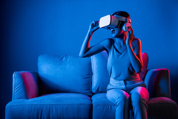 Smart female sitting on sofa wearing VR headset connecting metaverse, future cyberspace community technology. Elegant woman enjoy gesticulate interacting another users in virtual world. Hallucination. - obrazy, fototapety, plakaty