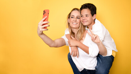 Happy young woman have fun child baby boy 12 years old in violet t-shirt do selfie shot on mobile phone. Mommy and son together isolated on yellow background