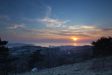 vista con inquadratura panoramica, dall'alto, sulla città di Trieste, il golfo ed il Mare Adriatico e i suoi dintorni, guardando verso sud-est, sotto un cielo parzialmente nuvoloso, al tramonto