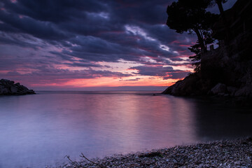 bellissimo tramonto in un cielo nuvoloso, con sfumature tra il rosso, il viola e il blu, che si riflette sul Mare Adriatico, visto dall'area marina della costiera vicino a Trieste - obrazy, fototapety, plakaty