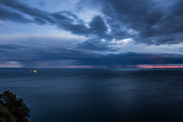 inquadratura panoramica dall'alto, vicino a Sistiana che mostra un temporale lontano che si abbatte sul Golfo di Trieste, nel Mare Adriatico, di sera, al tramonto, con un cielo completamente nuvoloso, - obrazy, fototapety, plakaty
