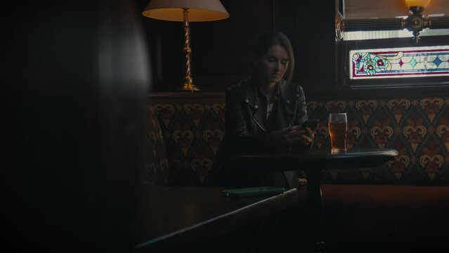 Young woman sitting in a pub, with a drink, texting on here phone, waiting. 