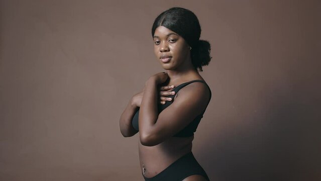 Young attractive African American woman in lingerie touching skin lovingly and hugging herself while posing for camera on coffee color cyc wall in studio