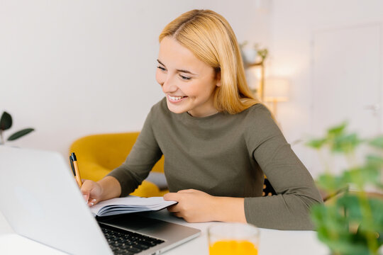 Young student woman taking notes while using laptop computer at home. Millennial european female learning online listening virtual video call. Business and education concept.