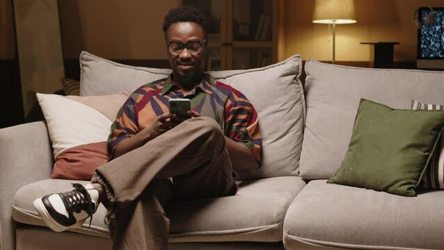 Medium shot of young Black hipster man in colorful printed shirt sitting on couch in modern living room and scrolling on smartphone in afternoon