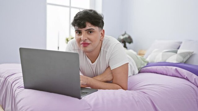 Young man with styled hair smiling while using laptop on purple bed in bright bedroom
