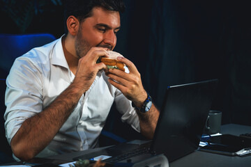 Businessman enjoy eating with delicious sandwich dish on desk while staring laptop at night time....