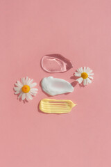 Smears of yellow, white cream and transparent gel or serum with chamomile flowers on a pink background. The texture of a cosmetic skin care product. The concept of natural cosmetics. Top view.