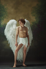Young man, with cascading curly hair and colossal white wings, stands exudes serenity against vintage studio background. Concept of fashion and beauty, modern art and historical fiction fusion. Ad