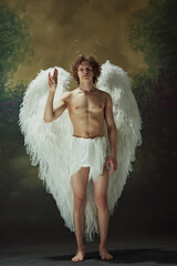 Curly-haired man posing looks like angel, with white big wings showing peace sign against vintage studio background. Concept of fashion and beauty, modern art and historical fiction fusion. Ad