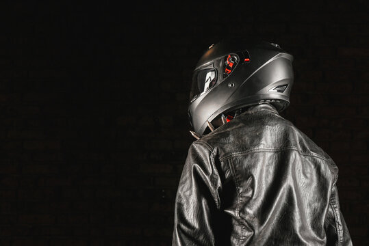 Brutal motorcyclist rider in black helmet and black studded leather jacket on a dark background.
