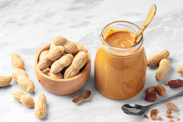 Peanut butter in glass jar and peanuts in wooden bowl on marble table