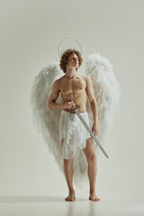 Man with white wings looks like angel holds sword against white studio background, his presence with sense of divinity. Concept of fashion and beauty, modern art and historical fiction fusion. Ad