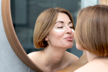 Attractive woman looking at mirror and applying moisturizing cream on cheeks in bathroom. Groomed...