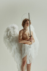 Man holds sword, his wings spread wide like angel, model looks to side embodying valor and grace against white studio background. Concept of fashion, beauty, modern art and historical fiction fusion.