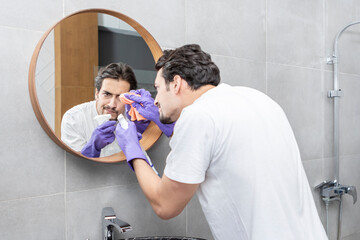 Handsome man cleaning mirror in the bathroom. Male contractor from cleaning service i yellow...