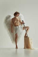 Young man with large wings posing holds sword exuding confidence and valor against white studio background. Concept of fashion and beauty, peace, modern art and historical fiction fusion. Ad