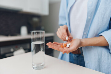 Woman holding pills and a glass of water on countertop in a modern kitchen interior with natural...